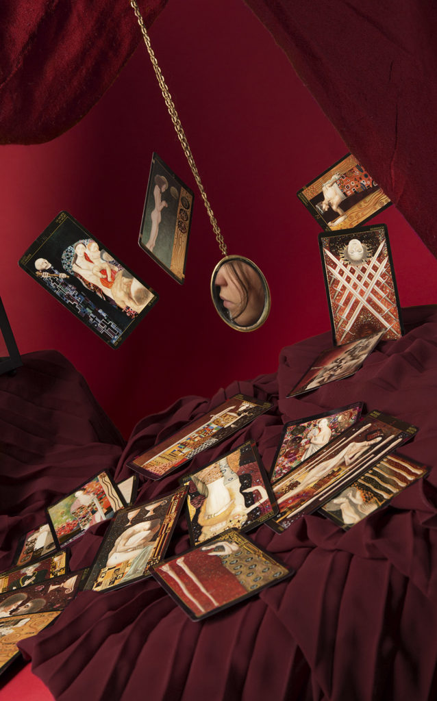 An image depicting a tabletop with tarot cards swirling above it against a red background, a small mirror shows a glimpse of a face